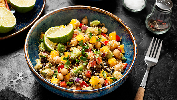This is an image of a blue ceramic bowl containing cooked quinoa, chickpeas, diced red peppers, diced red onion, black beans, cubed mango, cubed avocado, and sliced lime.